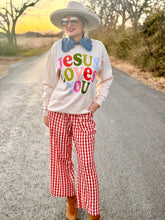 Person wearing a colorful 'Jesus Loves You' shirt, red checkered pants, and a wide-brimmed hat on a road.