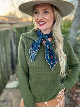 Woman wearing a green sweater and beige hat with a blue scarf, standing outdoors.